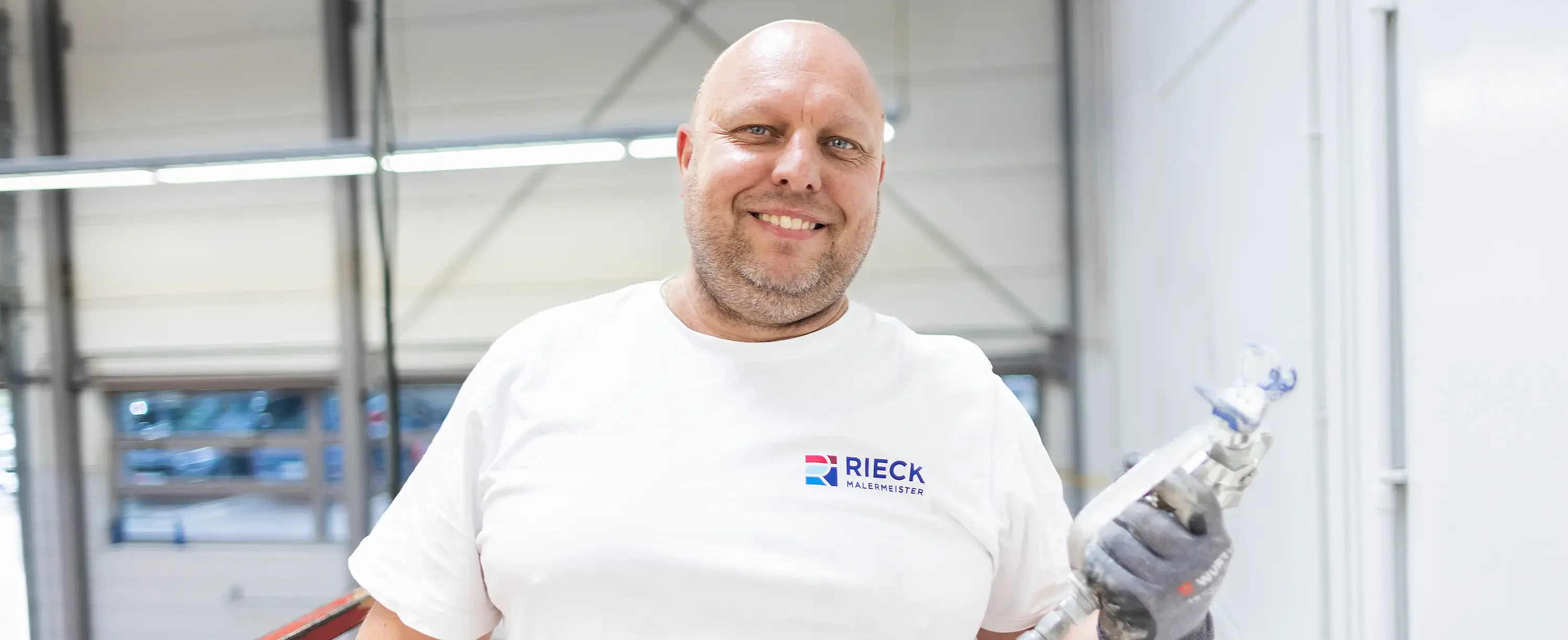 Handwerker mit Spritzpistole steht auf Arbeitsbühne im Innenraum bei Malerarbeiten.