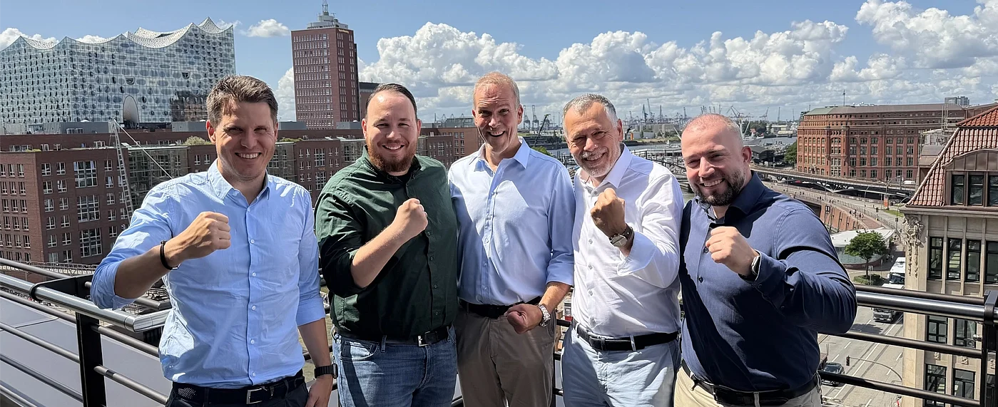 Fünf Männer schauen lächelnd in die Kamera machen die "wir packen an Geste" im Hintergrund die Skyline der Hamburger Hafen City 