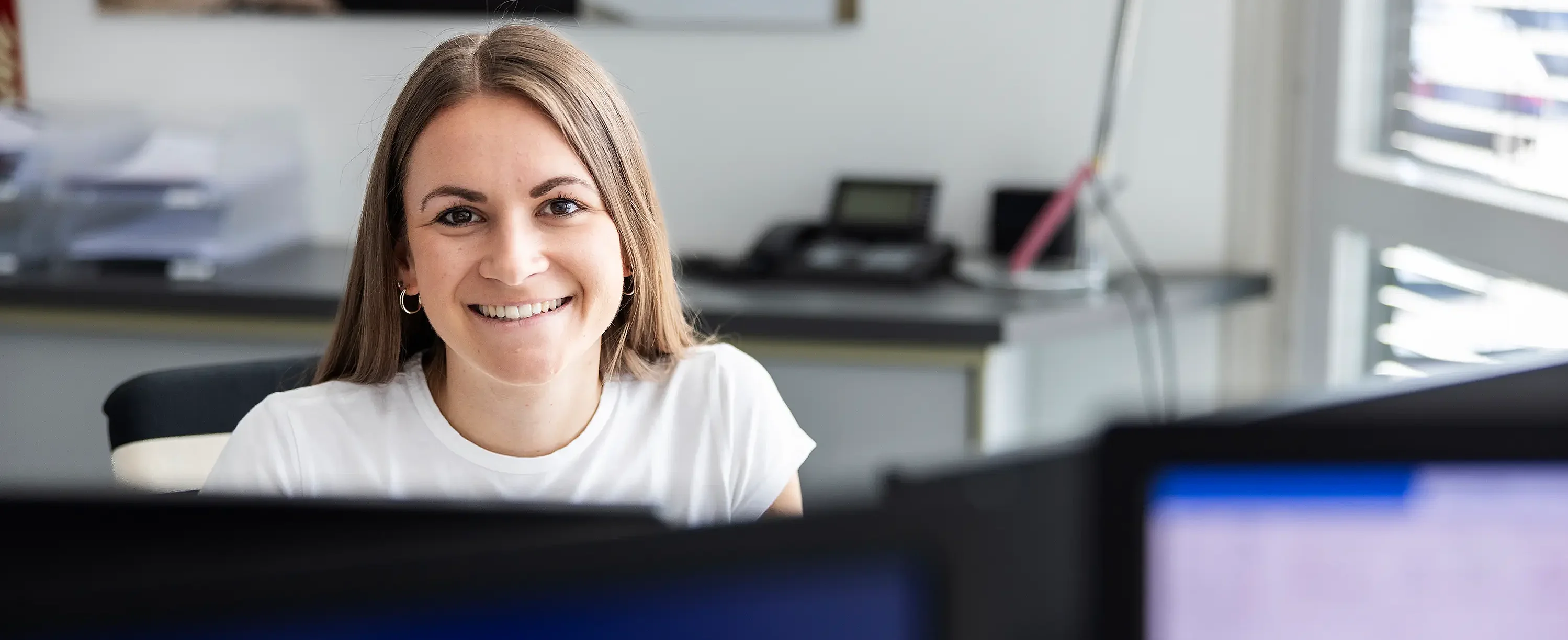 Mitarbeiterin im Büro sitzt am Schreibtisch und arbeitet vor mehreren Monitoren im Arbeitsumfeld.