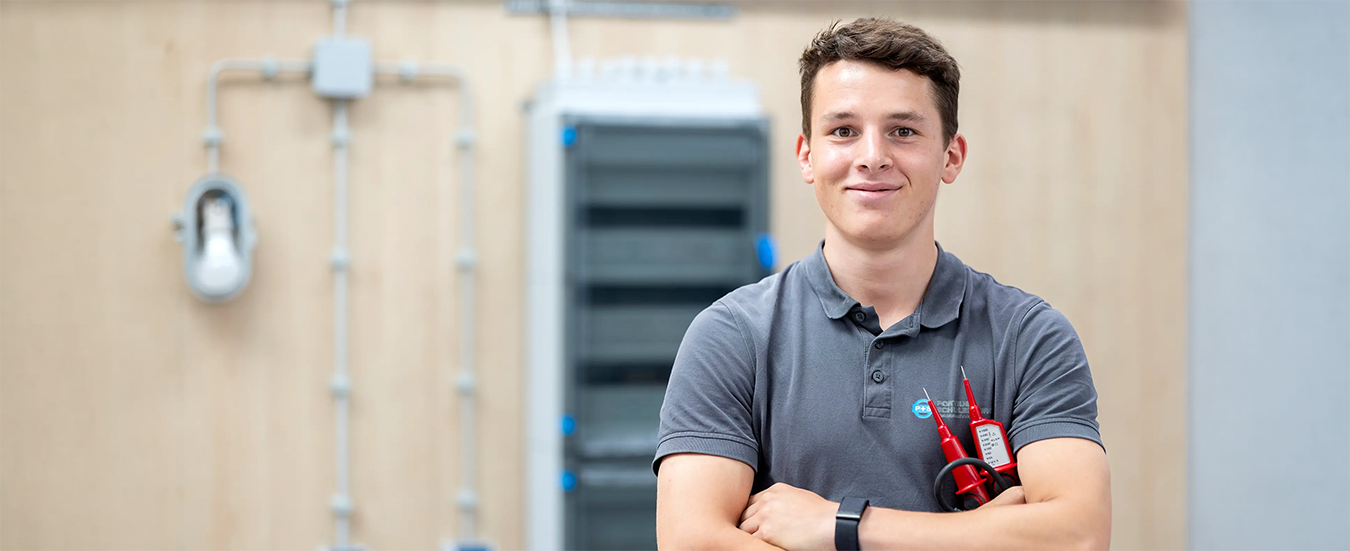 Auszubildender im Elektrohandwerk hält Messgerät in Werkstatt mit Schaltanlagen im Hintergrund.