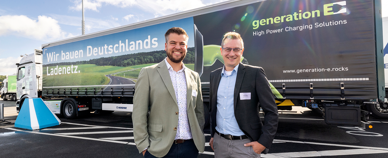 Zwei Geschäftspartner präsentieren vor einem Generation-E-Lkw die Ladeinfrastruktur für elektrische Nutzfahrzeuge.